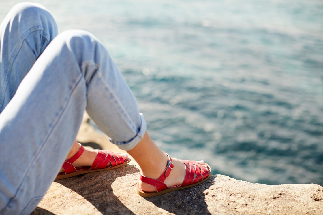 a woman is sitting on a wall, wearing red sandals, overlooking the sea