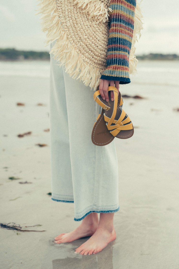 Woman on a beach, with her feet in the wet sand is holding a pair of mustard yellow, flat, woven leather sandals